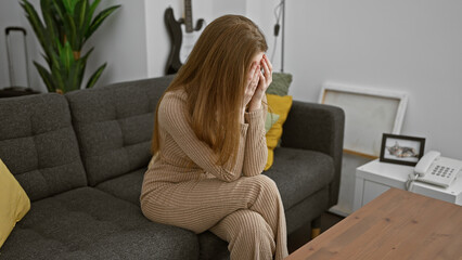 Obraz premium A distraught young woman sitting in a modern living room, covering her face with her hands