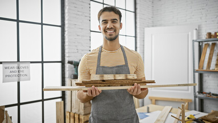 Smiling young hispanic man with a beard posing in a workshop holding wood.