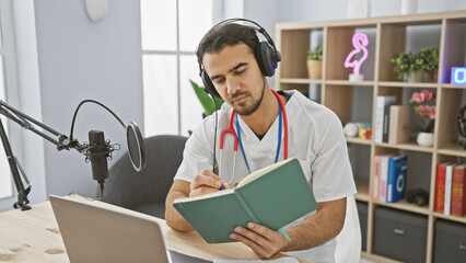 A handsome young man in headphones and a lab coat reads a book near a microphone in a podcast...