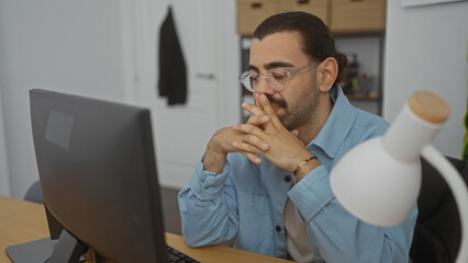 Young man with glasses and moustache thoughtfully working at computer in a modern office setting