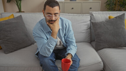 Young man with moustache holding red mug sitting in living room, wearing glasses and blue jacket, appearing thoughtful