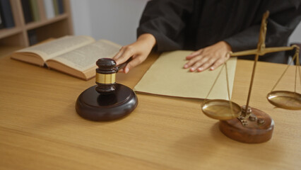 Woman judge sitting at a desk in a courtroom with scales of justice, a gavel, and law books, emphasizing her hands and the professional office environment