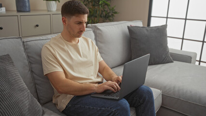 Handsome young hispanic man working on a laptop while sitting on a couch in a modern living room at home.