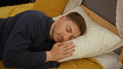 A young, attractive man sleeps peacefully in a cozy bedroom, lying on a bed with a yellow blanket and white pillow indoors.