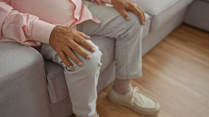 An elderly man sits indoors on a sofa in a living room with his hand resting on his knee.