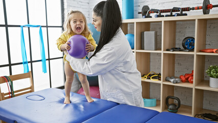 A pediatrician assists a young girl with physiotherapy exercises using a ball in a clinic's rehab room.