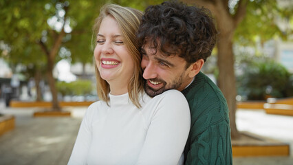 A happy couple embraces in a sunny park with lush green trees in the background.