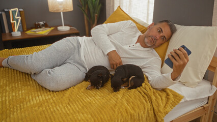 Middle-aged hispanic man relaxing on bed with two chihuahua dogs in a cozy bedroom, using a smartphone.