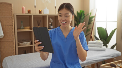 Asian woman in a blue uniform having a video call in a serene spa room with a massage bed and candles, conveying a sense of wellness and relaxation.