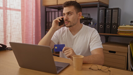 Young hispanic man talking on phone in office holding credit card, working on laptop, and having coffee.