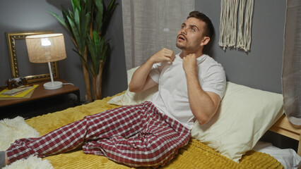 Handsome hispanic man with a beard sits on a bed in a cozy bedroom, dressed in plaid pajamas,...
