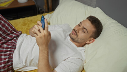 Handsome young hispanic man with a beard lying in bed in a bedroom at home, using a smartphone while wearing a white shirt and plaid pajama pants.