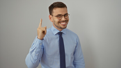 Handsome young hispanic man with a beard and glasses wearing a blue shirt and tie, smiling and pointing upwards, isolated on a white background.