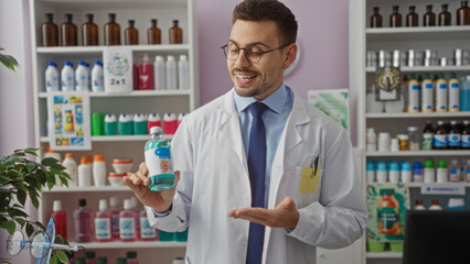 Fototapeta premium A handsome, young hispanic man with a beard wearing glasses and a white lab coat holds a product and smiles inside a pharmacy store with shelves full of various items.