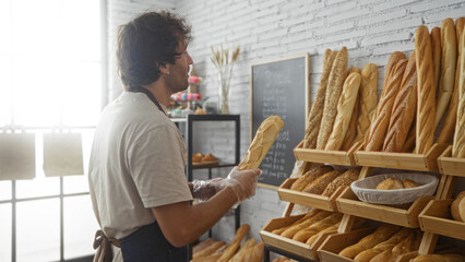 Young man choosing bread in a bakery filled with various types of baguettes and pastries, surrounded by wooden shelves and a chalkboard menu.