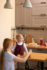Mum feeds her young daughter in the kitchen. The little girl sits on the table with a mouthful of food and smiles.