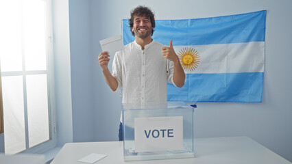 Young man in electoral room with argentinian flag giving thumbs up while holding ballot paper by a...
