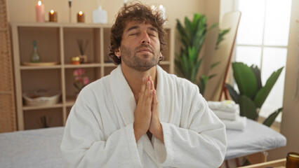 Young man in a white robe meditating in a serene spa room with candles and plants, exuding relaxation and wellness.