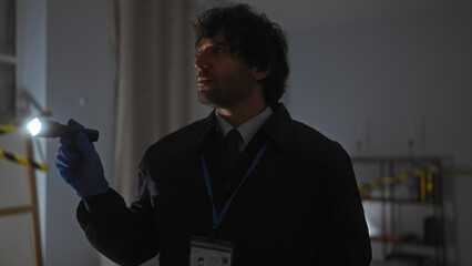 A handsome hispanic man intently searches a dark indoor crime scene with a flashlight and badge visible.