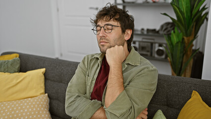 Obraz premium Pensive young hispanic man with beard and glasses sitting on a couch indoors with closed eyes