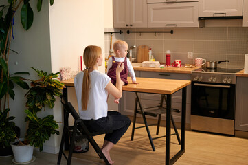 Mother feeding her baby. General view of the kitchen, mum sitting on a chair, little daughter on the table.