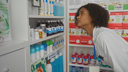 A young african american woman with curly hair examines shelves of medication in a pharmacy store interior.