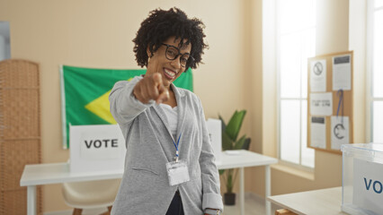 A beautiful young african american woman with curly hair stands in an electoral college room in brazil, smiling and pointing at the camera with a voting banner in the background.
