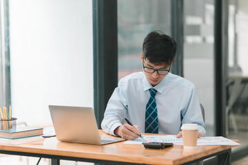A focused professional writes notes at a desk, using a laptop amidst a modern office environment.
