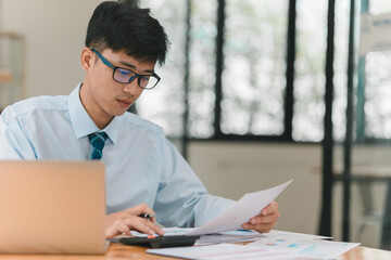 A focused individual in a blue shirt and glasses reviews documents while working at a desk with a laptop, showcasing a productive office environment.