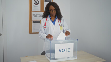 An african american woman with curly hair wearing a lab coat and stethoscope is voting indoors at an electoral college.