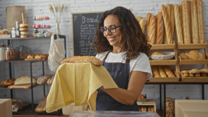 Woman holding freshly baked bread in a bakery room, with shelves of assorted bread and pastries in the background, wearing an apron and smiling, indoor setting in a small shop.