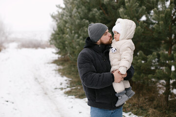 Bearded man is holding a baby dressed warmly in a white overall and hat, surrounded by snow-covered ground and evergreen trees. Joyful moment between the father and little daughter in a winter