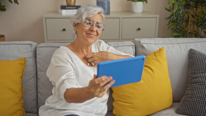 Obraz premium Elderly woman smiling while using a tablet in a cozy living room, sitting on a sofa with yellow pillows, showcasing a relaxed and comfortable home environment.