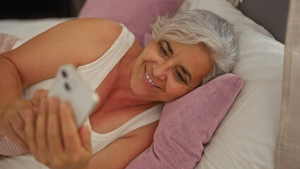 Elderly grey-haired woman lying on bed in a cozy bedroom, smiling while using a smartphone, capturing a moment of relaxation and joy indoors at home.