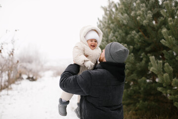 Bearded man is holding a smiling baby dressed warmly in a white overall and hat, surrounded by snow-covered ground and evergreen trees. Joyful moment between the father and little daughter in a winter