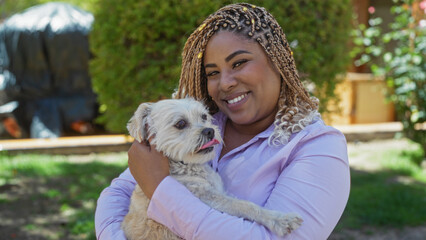 A young african american woman with braids smiles while holding her dog in an urban park on a sunny day.