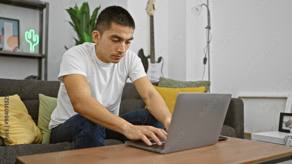 Wall mural a focused hispanic man using a laptop in a modern living room setup, epitomizing casual remote work.