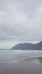 Scenic view of famara beach with waves crashing against the sandy shore under an overcast sky in lanzarote, canary islands, alongside a mountainous coastline.