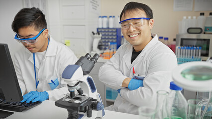 Two asian men in white coats working in a science lab, one using a computer, the other smiling with crossed arms.