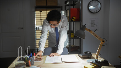 Asian man in office analyzing documents at night, portraying investigation work in a professional setting.