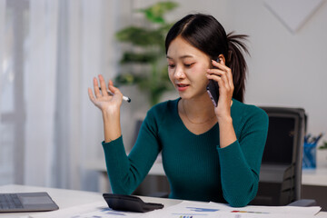 Asian young businesswoman talking on mobile phone and discussing business issue while working at laptop in modern office
