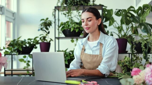 Florist small business owner wearing an apron stands working in her store surrounded by plants and flowers. She is smiling and looking at the camera while working on her laptop.