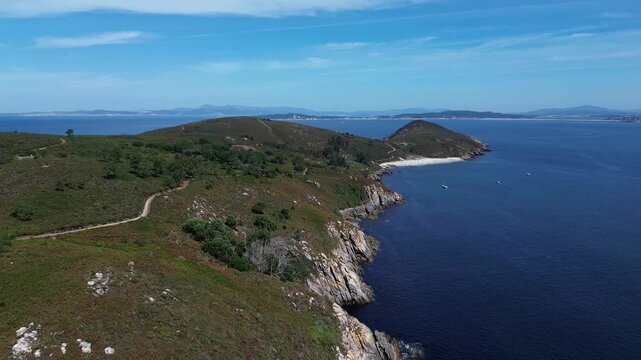 Overlooking the paths and rugged cliffs of Ons Island, the serene sea glistens. Melide Beach and anchored boats appear ahead, with Pontevedra&rsquo;s estuary in the distance under blue sky with high clouds.