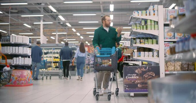 Caucasian Man Wearing Glasses and a Green Shirt Shops in a Modern Supermarket, Examining Canned Beans while Pushing a Cart. Shelves Are Stocked With Diverse Products and Shoppers Navigate the Aisles