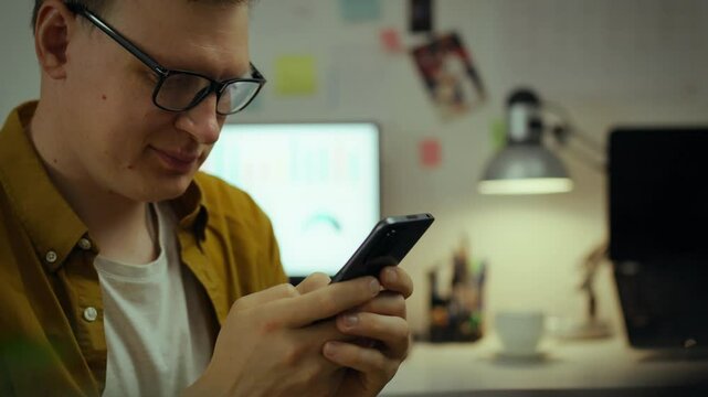 A future techie takes a break from classes, using a mobile gadget in dorm room. Next to the computer, young college student sits, interestedly studying the information on the screen of his smartphone