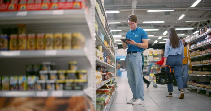 Caucasian Male Supermarket Manager Making an Inventory Review with the Help of a Tablet Computer. Young Worker Replenishing and Organizing Stock in an Aisle with Canned Food