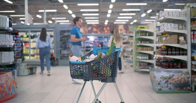Establishing Footage with a Shopping Cart Standing Between the Aisles in a Modern Grocery Store. Busy Supermarket with Diverse People in the Background. Trolley with Vegetables, Milk, Canned Foods