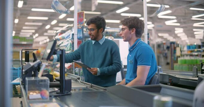 Young Indian Supermarket Manager Having a Conversation with a Cashier. Colleagues Discussing New Store Checkout System and Watching Instructions in a Demo on a Tablet Computer