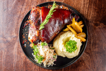 succulent roasted pork knuckle garnished with fresh herbs, served with mashed potatoes and sauerkraut on black plate against wooden backdrop. Perfect for culinary presentations and food photography.