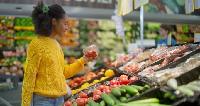 Beautiful Young Black Woman Shopping for Fresh Produce in a Modern Supermarket, Standing at a Vegetable Aisle and Carefully Selecting Tomatoes and Other Items From the Neatly Arranged Shelves